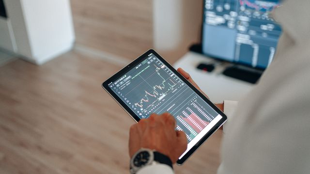 A close-up of a person holding a tablet displaying financial stock market charts and candlestick graphs, with a trading screen blurred in the background.