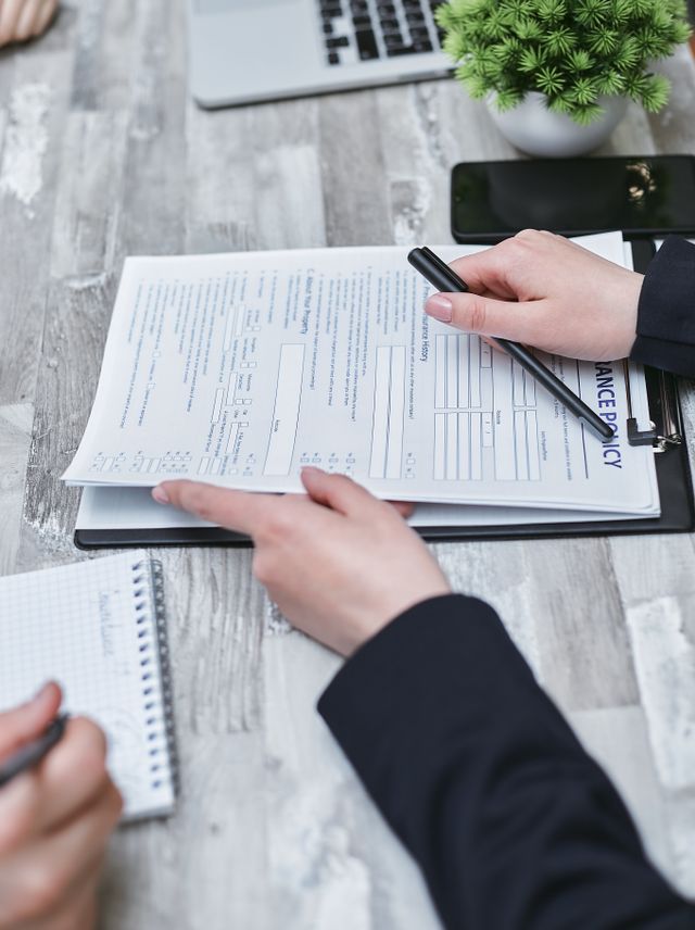 A high-angle, close-up shot of hands reviewing an insurance policy document on a clipboard. One person in a dark blazer holds a black pen, while another person's hand stabilizes the paperwork on a wooden table setup with a laptop, smartphone, and small green plant in the background.