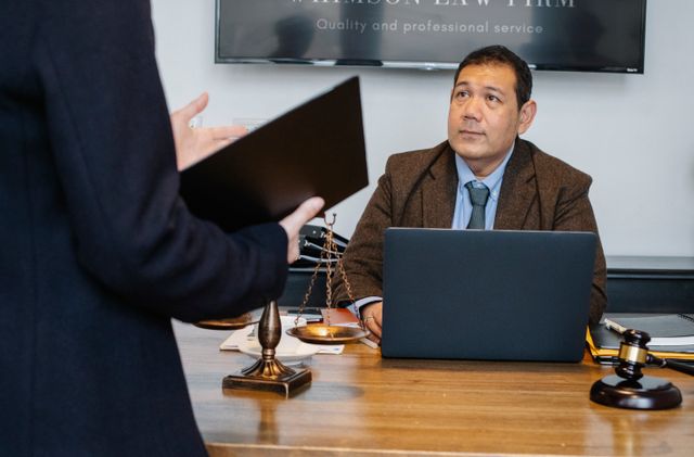 "An immigration lawyer sitting at his office desk with a laptop, scales of justice, and a gavel, listening attentively to a client presenting case documents."