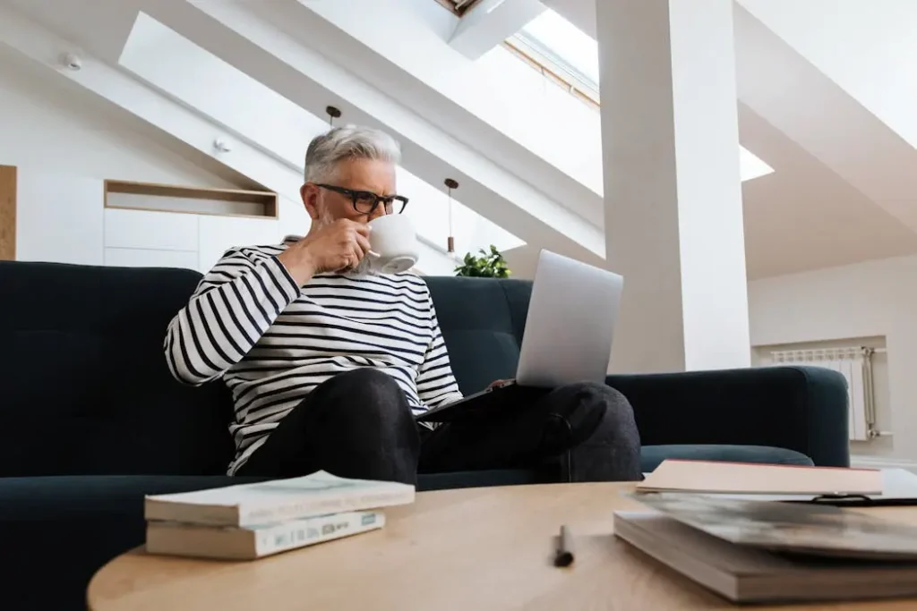 A mature investor relaxing on a sofa with coffee while using a laptop to manage his self directed ira portfolio.