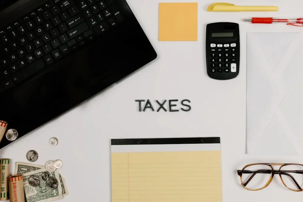 A flat lay of a desk with a laptop, calculator, cash, and wooden tiles spelling TAXES, representing retirement and dividend tax planning.