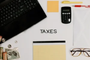 A flat lay of a desk with a laptop, calculator, cash, and wooden tiles spelling TAXES, representing retirement and dividend tax planning.
