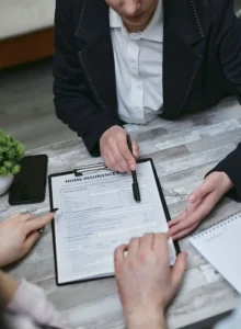 A close-up shot of an insurance agent holding a pen and pointing at a "Home Insurance Policy" form on a clipboard, explaining the coverage details to a couple sitting across the wooden table.