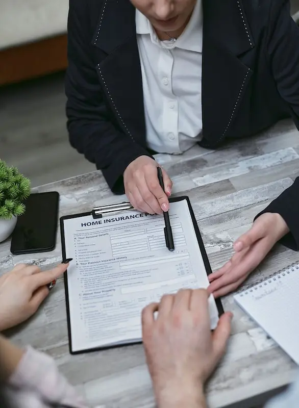 A close-up shot of an insurance agent holding a pen and pointing at a "Home Insurance Policy" form on a clipboard, explaining the coverage details to a couple sitting across the wooden table.