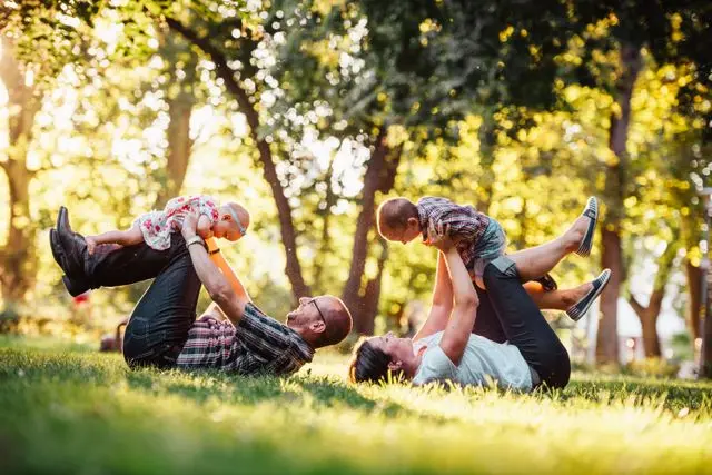 A happy family with a mother, father, and two young children playing on the grass in a sunny park, representing long-term financial security and life insurance protection.