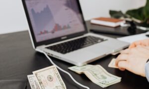 A laptop displaying financial market charts on a desk with US dollar bills, representing passive income investments.