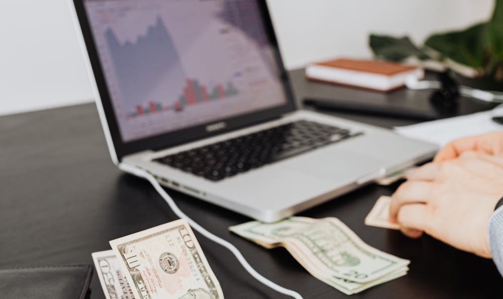 A laptop displaying financial market charts on a desk with US dollar bills, representing passive income investments.