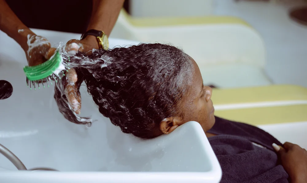 A Black woman receiving a chemical hair treatment in a salon basin, representing the potential chemical exposure linked to uterine and ovarian cancer claims in hair relaxer lawsuits.