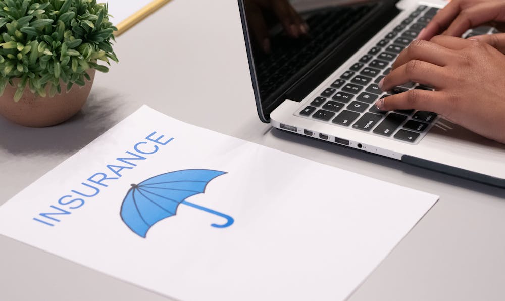 A person typing on a laptop next to a document featuring the word "Insurance" and a blue umbrella icon, representing research on professional liability insurance cost and coverage to protect a business.