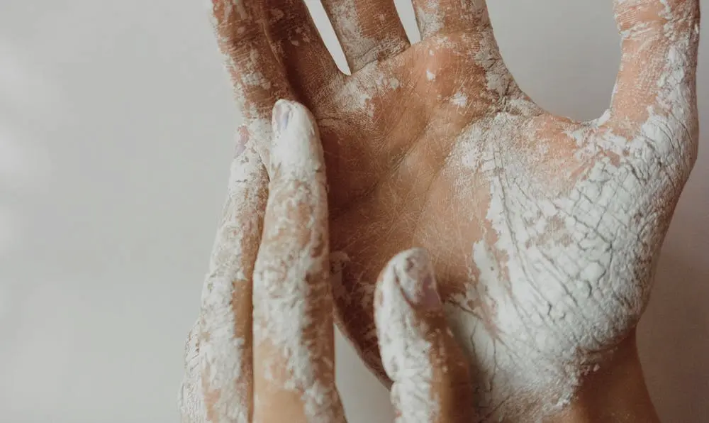 "A close-up shot of hands coated in a fine, dry white powder. The substance exhibits a distinctive cracked texture on the skin, evocative of the heavy application of talcum or baby powder."