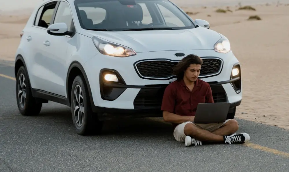 Young man using a laptop next to a white SUV on a desert road.