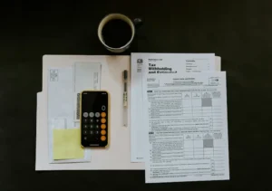 A top-down view of a desk featuring a smartphone calculator, a pen, and official IRS tax forms, representing the preparation needed to report digital payment income.