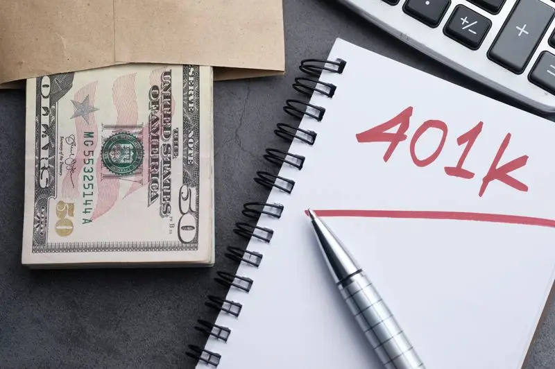 "A close-up top view of a spiral notepad with '401k' written in red ink next to a stack of fifty-dollar bills in an envelope, a silver pen, and a calculator, representing retirement planning and 2026 contribution limits."