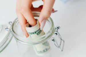 A close-up view of a hand placing a rolled stack of US dollar bills into a clear glass jar, concept of emergency fund and savings.