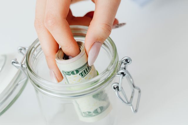 A close-up view of a hand placing a rolled stack of US dollar bills into a clear glass jar, concept of emergency fund and savings.