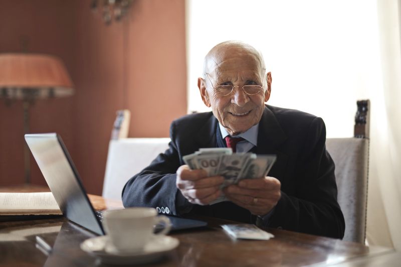 Smiling senior holding cash by his laptop, representing a well-funded retirement. (Average Retirement Savings by Age How You Compare.jpg)