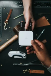 A person inserting a Capital One Visa credit card into a white Square contactless card reader to make a payment over a dark workbench filled with leathercraft tools.