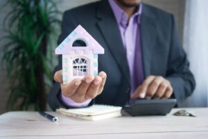 A real estate professional or financial advisor in a suit holding a small model house in one hand while using a calculator with the other, with a notebook and pen on the desk.