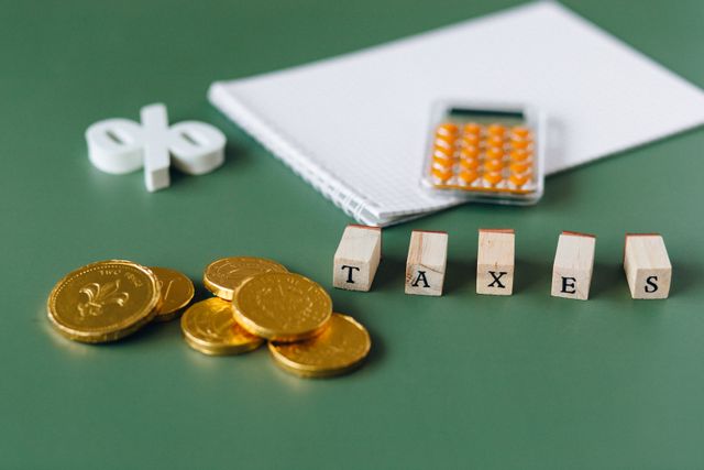 "A flat lay composition on a green background featuring wooden blocks that spell 'TAXES', surrounded by gold coins, a small orange calculator on a notepad, and a white percentage symbol."