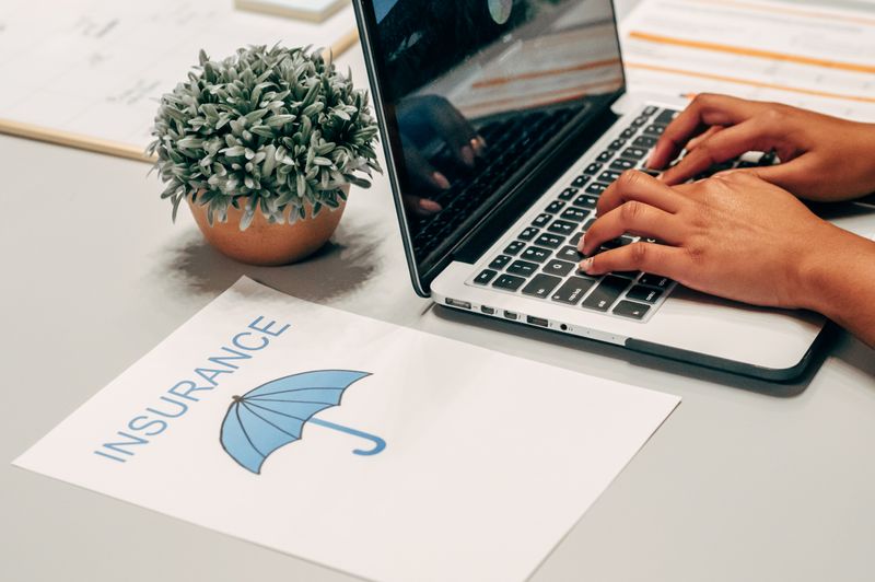 A close-up of hands typing on a laptop keyboard beside a printed document showing the word "INSURANCE" and a blue umbrella icon on a desk.