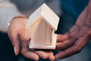 "Two people of different skin tones holding a small wooden model of a house together in their open palms, symbolizing partnership, protection, and homeownership."