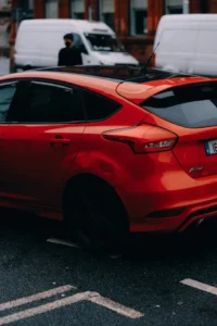 A red Ford Focus hatchback parked on a city street, featuring a black panoramic roof, tinted windows with rain guards, and distinctive rear taillights.