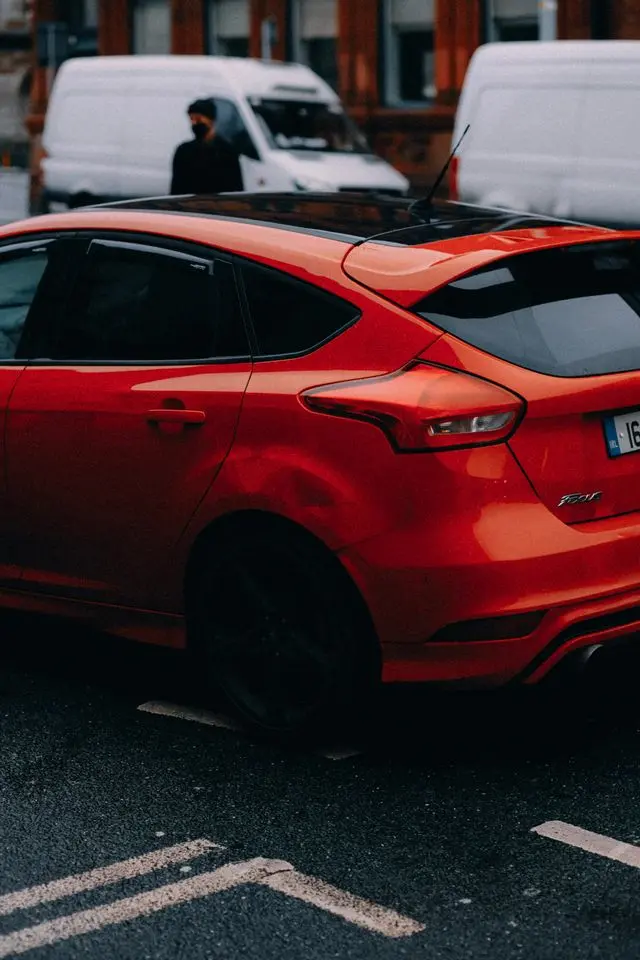 A red Ford Focus hatchback parked on a city street, featuring a black panoramic roof, tinted windows with rain guards, and distinctive rear taillights.