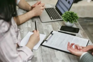 A close-up of people sitting at a desk, reviewing a printed insurance policy document on a clipboard next to a laptop, a notepad, and a smartphone.
