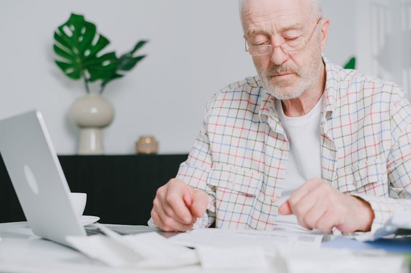 A senior man wearing glasses sitting at a desk with a laptop and paperwork, carefully reviewing documents to learn how to compare Medicare Advantage plans for seniors.