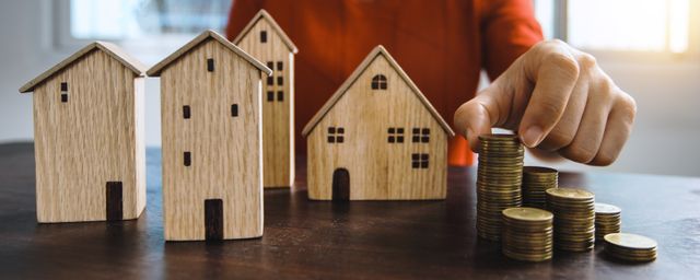 Wooden house models next to stacked gold coins on a table with a person's hand adding to the pile, illustrating how to invest in real estate in 2026.