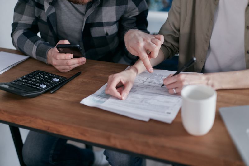 A close-up of a couple sitting at a wooden desk reviewing financial documents, with one person pointing at Form 1099-C while the other takes notes, next to a smartphone and a calculator.
