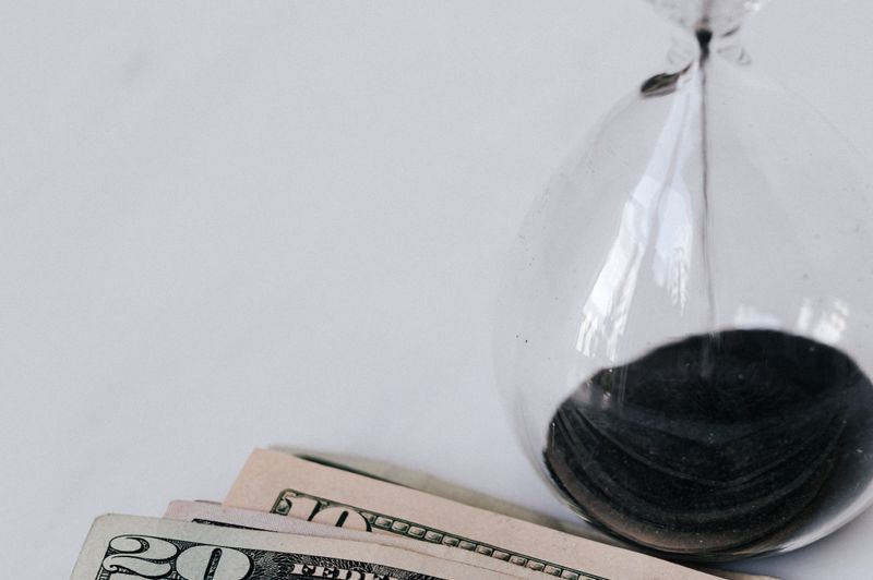 An hourglass with black sand flowing next to a stack of US dollar bills on a plain white background, symbolizing time, financial growth, and interest earnings.