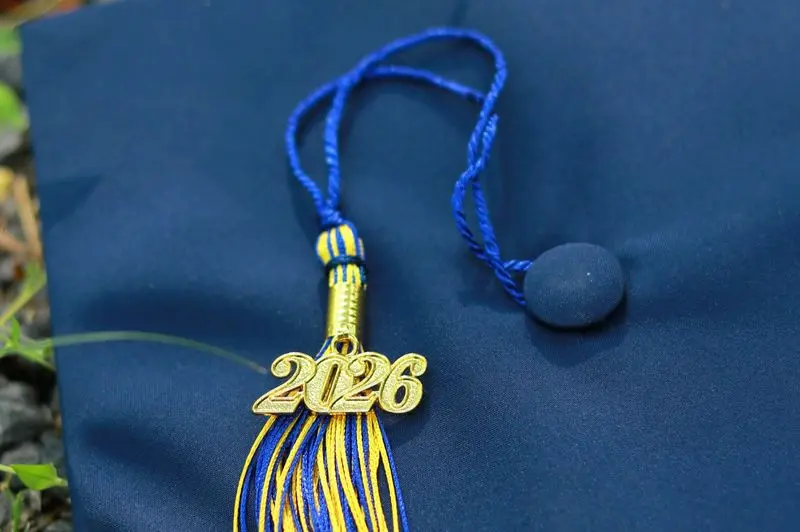A close-up of a dark blue graduation cap featuring a blue and gold tassel with a metallic gold "2026" year charm resting on top.