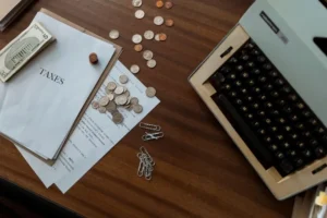 An administrative desk with a vintage typewriter on the right, tax documents labeled "TAXES" on the left, US dollar bills, scattered silver and copper coins, and paperclips spread across a dark wooden table.