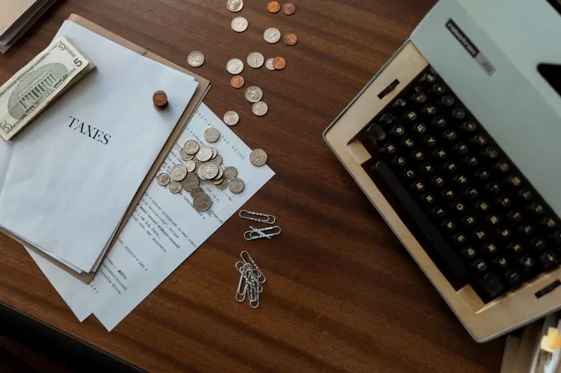 An administrative desk with a vintage typewriter on the right, tax documents labeled "TAXES" on the left, US dollar bills, scattered silver and copper coins, and paperclips spread across a dark wooden table.
