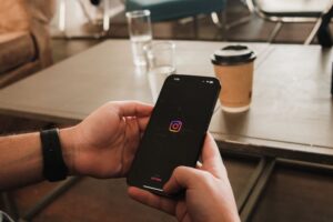 A person holding a smartphone that displays the Instagram launch screen with its colorful logo on a black background, set against a blurred coffee shop table.