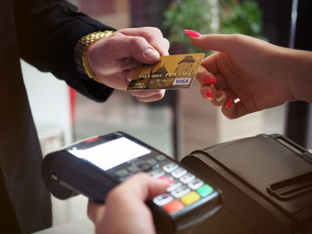 A close-up shot of a person’s hand passing a Visa credit card to another person over a black payment terminal machine at a checkout counter.