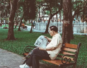 An elderly man sitting on a wooden bench in a shaded green park while reading a newspaper.