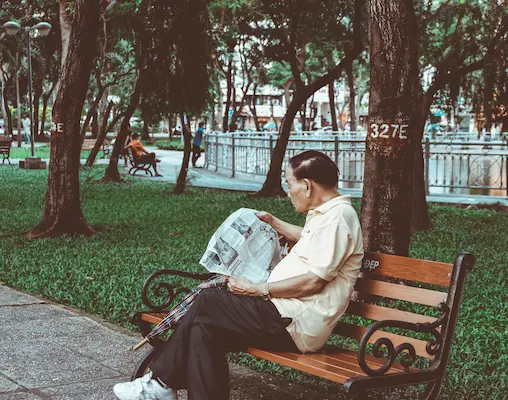 An elderly man sitting on a wooden bench in a shaded green park while reading a newspaper.