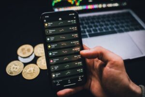 "Close-up of a hand holding a smartphone displaying a crypto trading application with a list of digital assets including Cardano (ADA), with physical crypto coins and a laptop in the background."