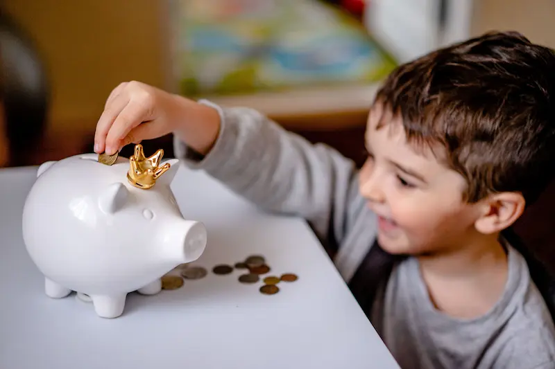 A smiling young boy saving coins in a white piggy bank with a gold crown, symbolizing wealth building and the 2026 Trump Accounts for children.