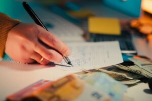 "A close-up of a person's hand signing financial documents with a pen, surrounded by cash notes and paperwork, symbolizing student loan applications, repayment planning, or financial management."