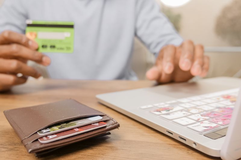 A person holding a credit card and using a laptop to manage finances and pay off debt online, with a wallet full of cards on a wooden desk.
