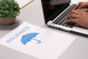 A person typing on a laptop next to a sheet of paper with the word "INSURANCE" and a blue umbrella graphic on a desk.