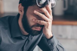 An image of a man looking stressed with a hand over his face. He is wearing a button-down shirt and has glasses resting on top of his head.