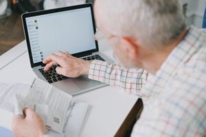An elderly man viewed from behind his shoulder, looking at an open laptop on a white desk while holding paper bills or financial documents in his hand, preparing to manage his finances.