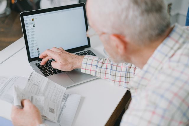 An elderly man viewed from behind his shoulder, looking at an open laptop on a white desk while holding paper bills or financial documents in his hand, preparing to manage his finances.