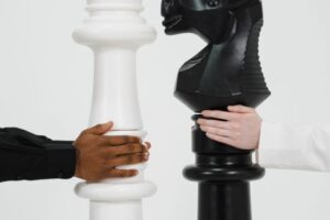 "Two giant chess pieces, a white king and a black knight, being held by two hands representing a strategic competition or financial matchup."