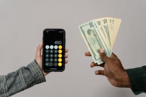 A hand holding a fan of US dollar bills next to another hand showing a calculator app with the number 450 on a smartphone screen.