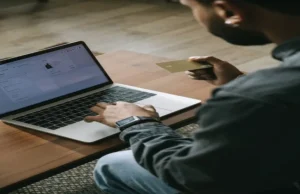 A man sitting at a desk, typing on his laptop while holding a gold credit card, analyzing personal vs business expenses on screen.